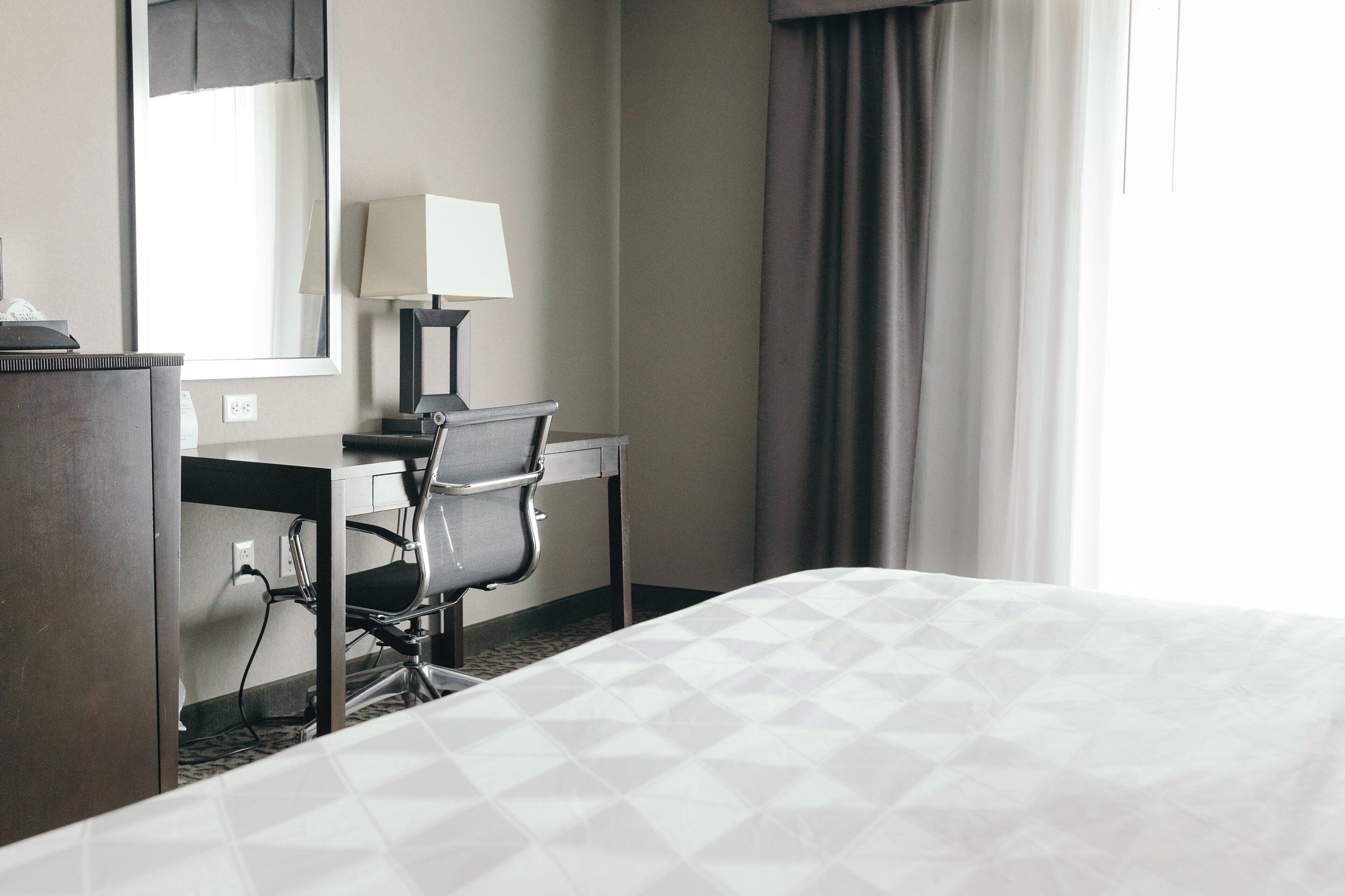 A hotel room with a bed, desk, chair, lamp, and a window with curtains.