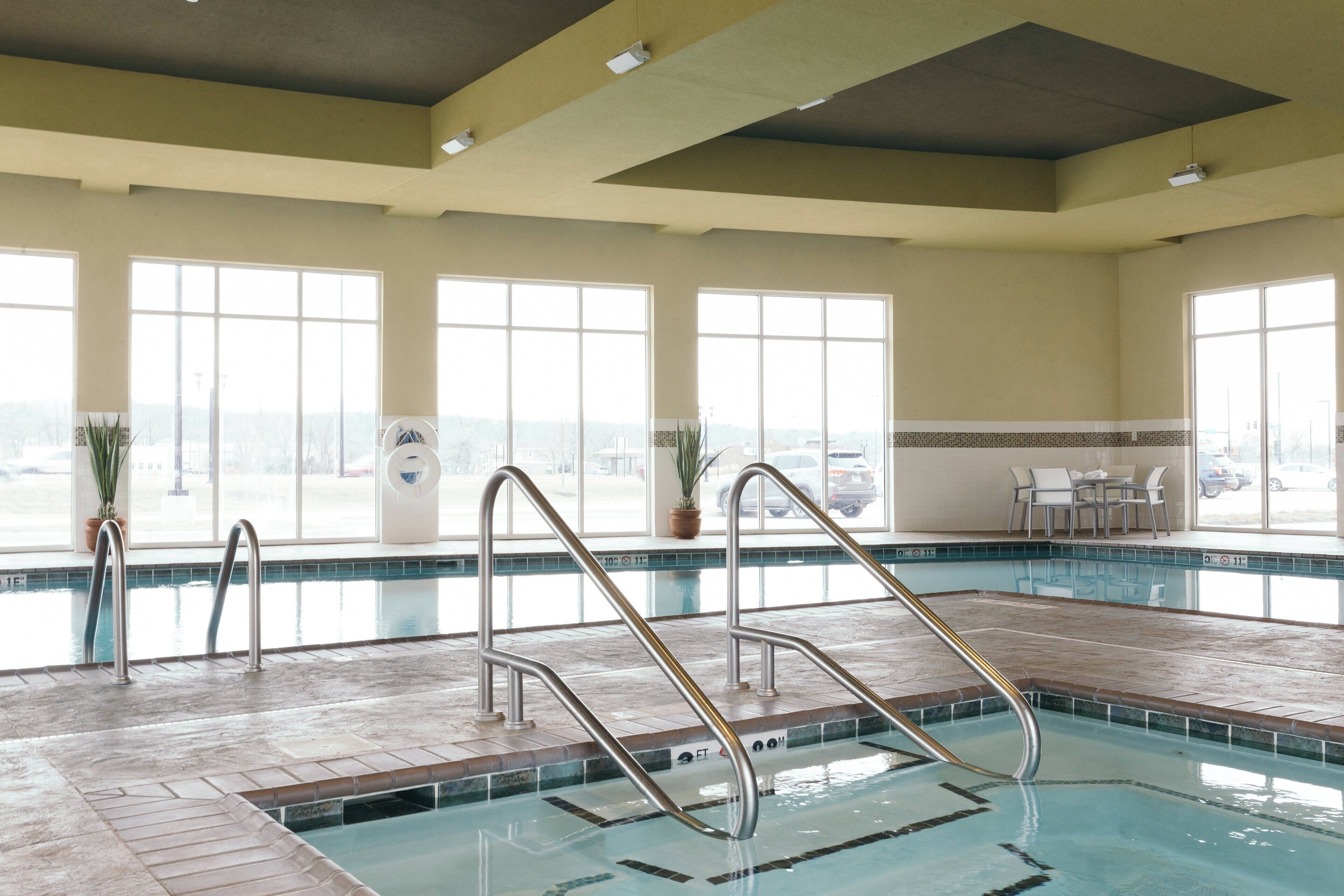 An indoor swimming pool with clear water, metal pool ladders, and a view of the city through large windows.