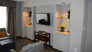 A modern living room with a flat-screen TV mounted on the wall, a sofa, a coffee table, and two shelves with potted plants and decorative items. - Holiday Inn Hotel & Suites East Peoria by IHG (East Peoria)