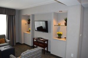 A modern living room with a flat-screen TV mounted on the wall, a sofa, a coffee table, and two shelves with potted plants and decorative items. - Holiday Inn Hotel & Suites East Peoria by IHG (East Peoria)