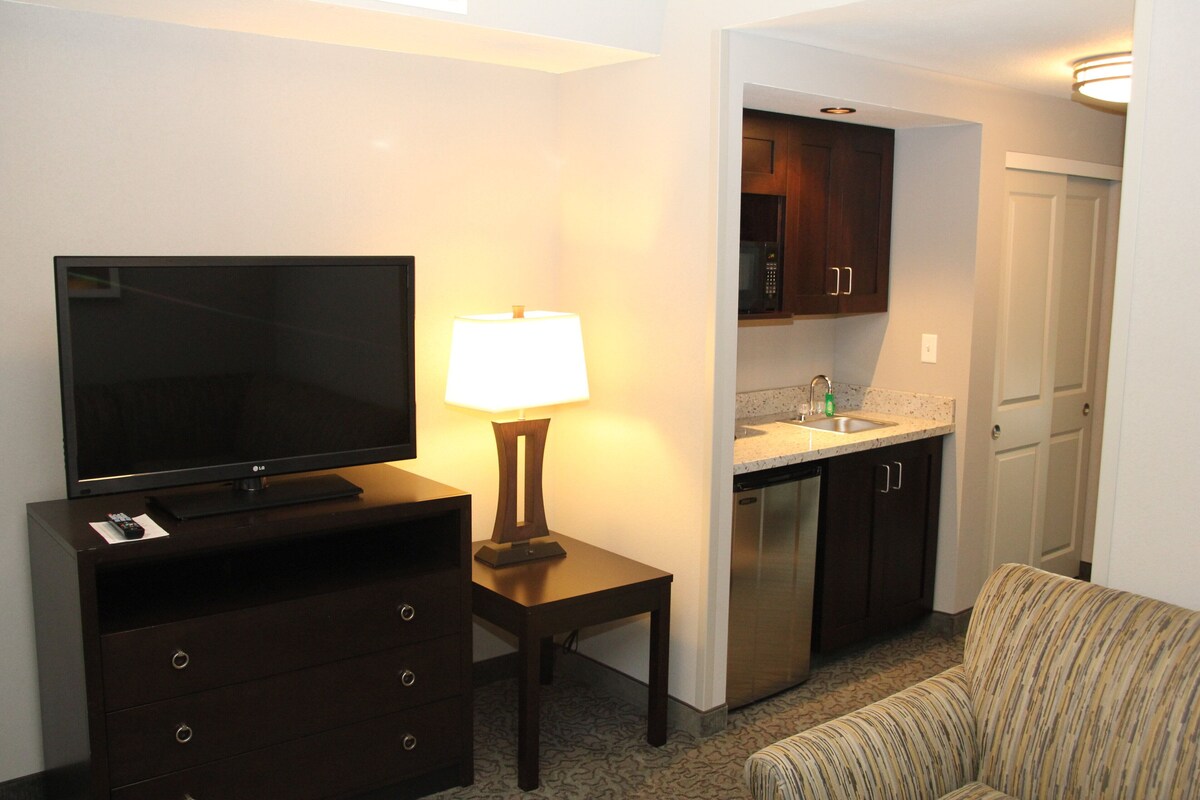 A compact hotel room with a TV, a lamp, a sofa, a kitchenette, and a door.