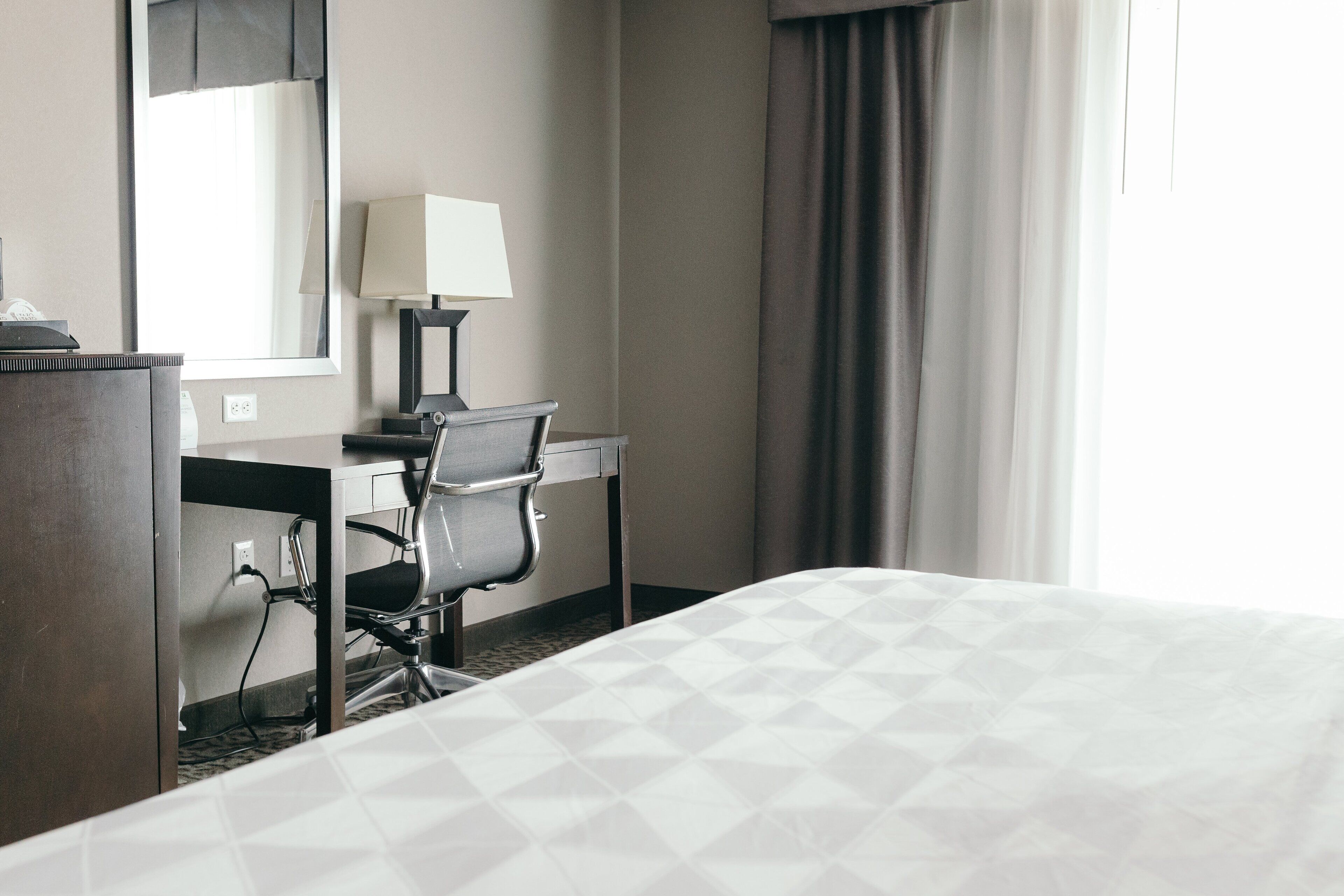 A hotel room with a bed, desk, chair, lamp, and a window with curtains.