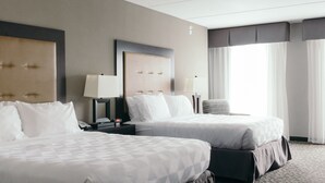 A hotel room with two beds, a nightstand with a lamp, a chair, and a large window with curtains. - Holiday Inn Hotel & Suites East Peoria by IHG (East Peoria)