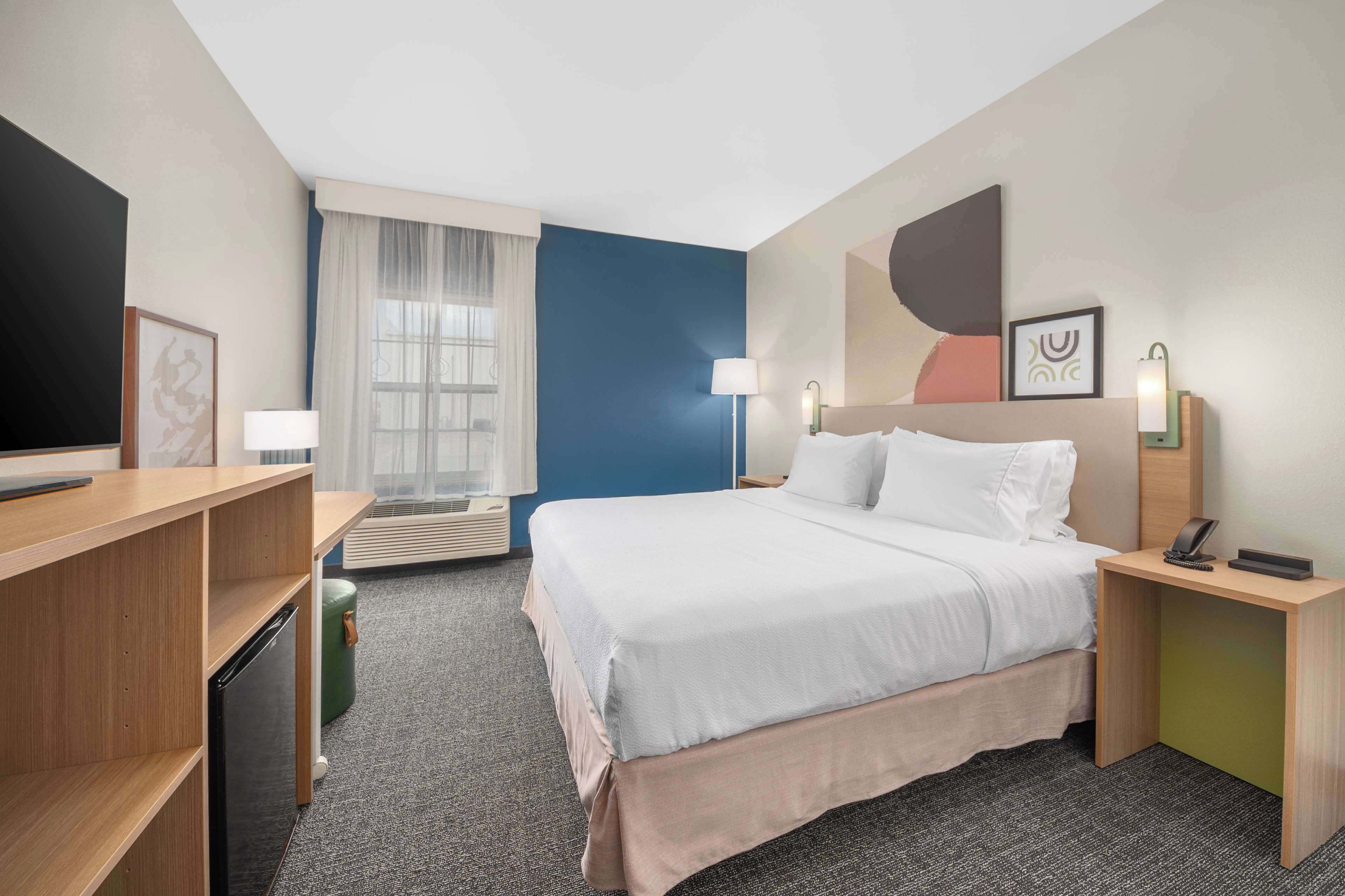 Una habitación de hotel con cama, mesitas de noche, escritorio, televisión y ventana con cortinas, además de un cuadro en la pared.
