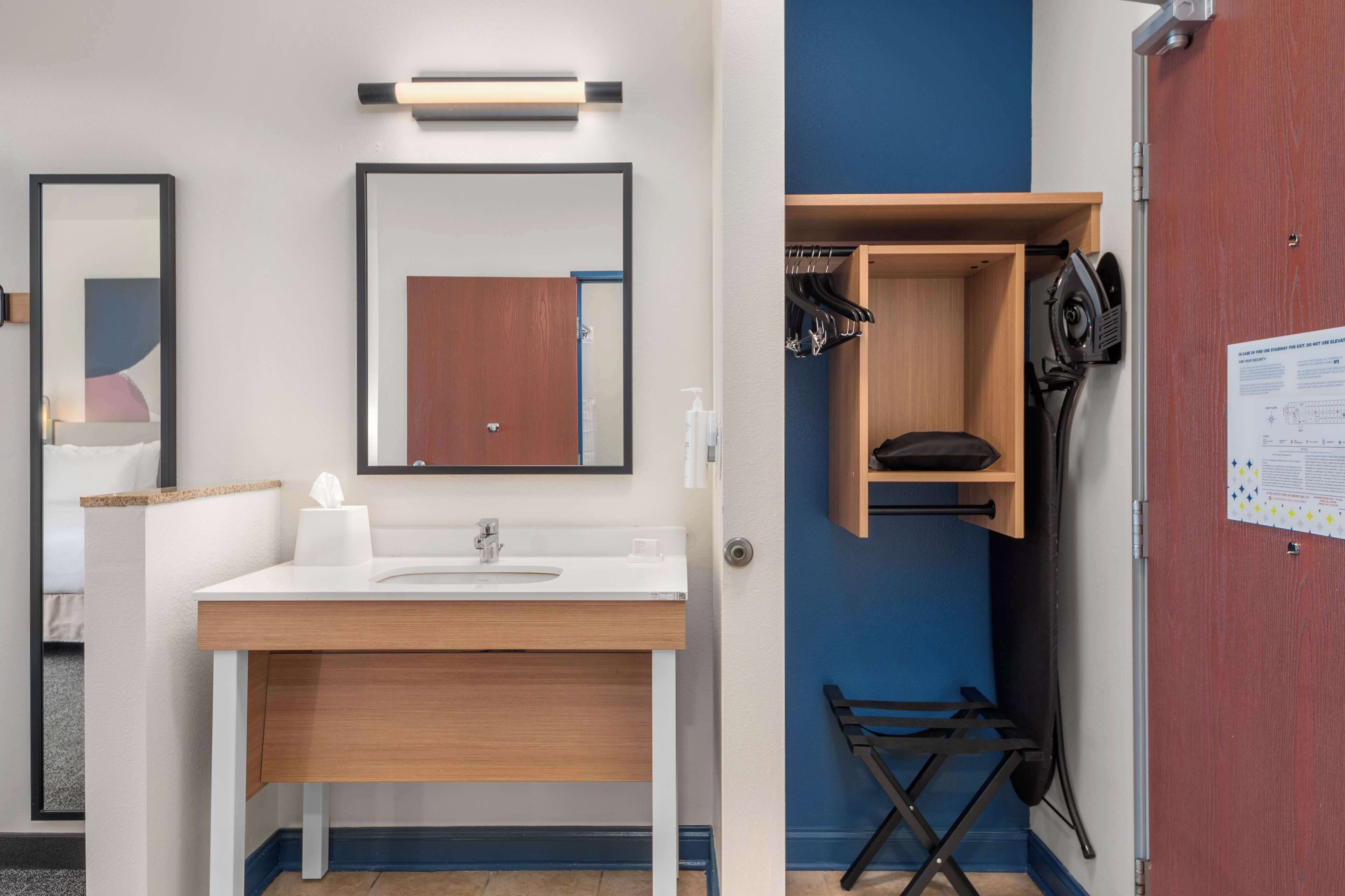 A modern bathroom with a white sink, wooden vanity, and a mirror. There is a blue wall and a wooden door with a coat rack and ironing board.