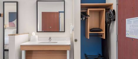 A modern bathroom with a white sink, wooden vanity, and a mirror. There is a blue wall and a wooden door with a coat rack and ironing board.