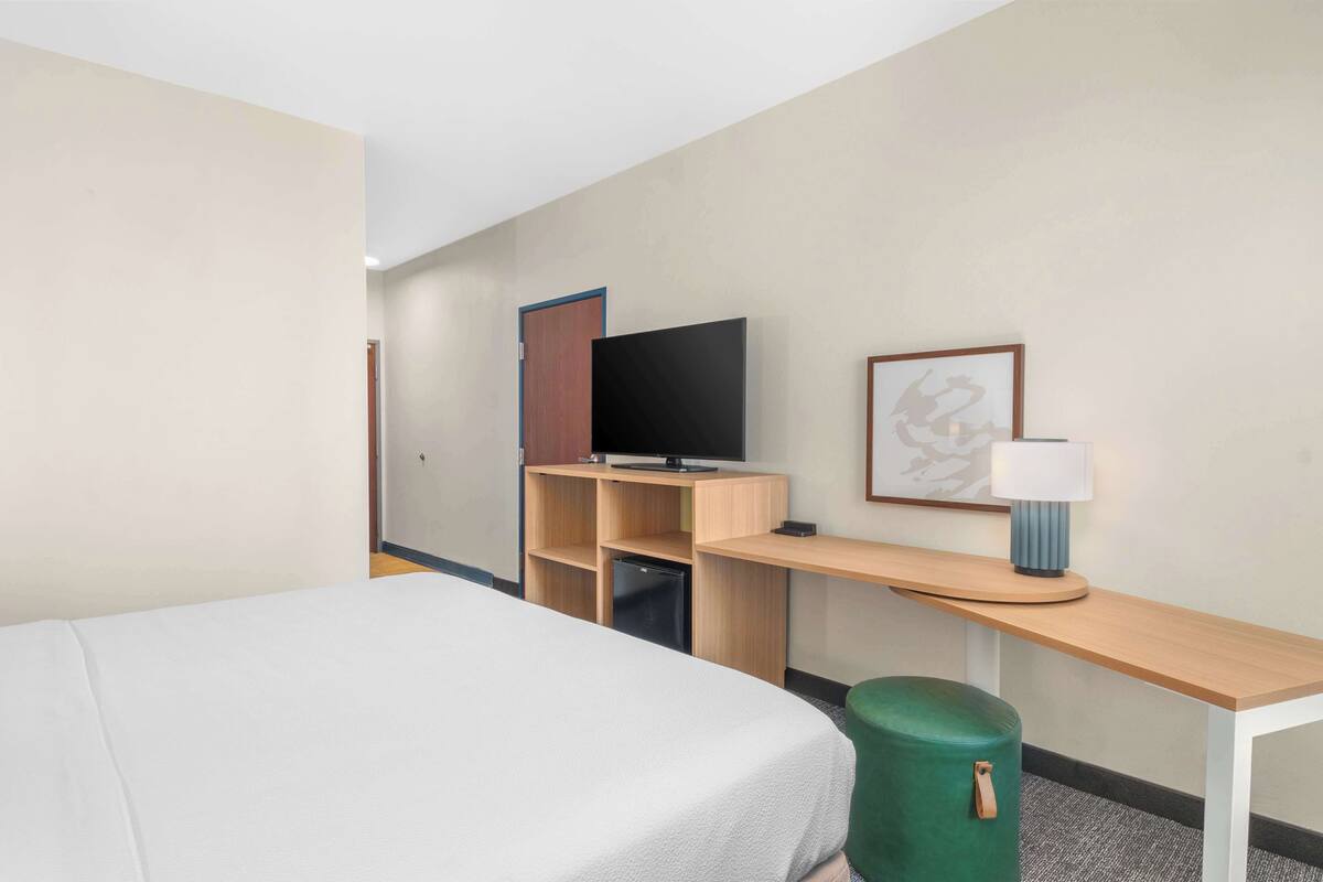 A hotel room with a bed, a desk with a television, a small refrigerator, and a green trash bin.