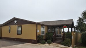 A single-story building with a brown and yellow exterior, a porch with a staircase, and a 'OPEN' sign. - Cotulla Homes (Cotulla)