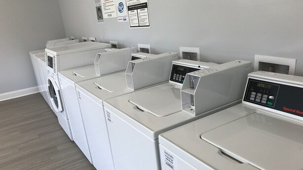A laundry room with multiple white washing machines and dryers, a wooden floor, and informational posters on the wall. - Cotulla Homes (Cotulla)