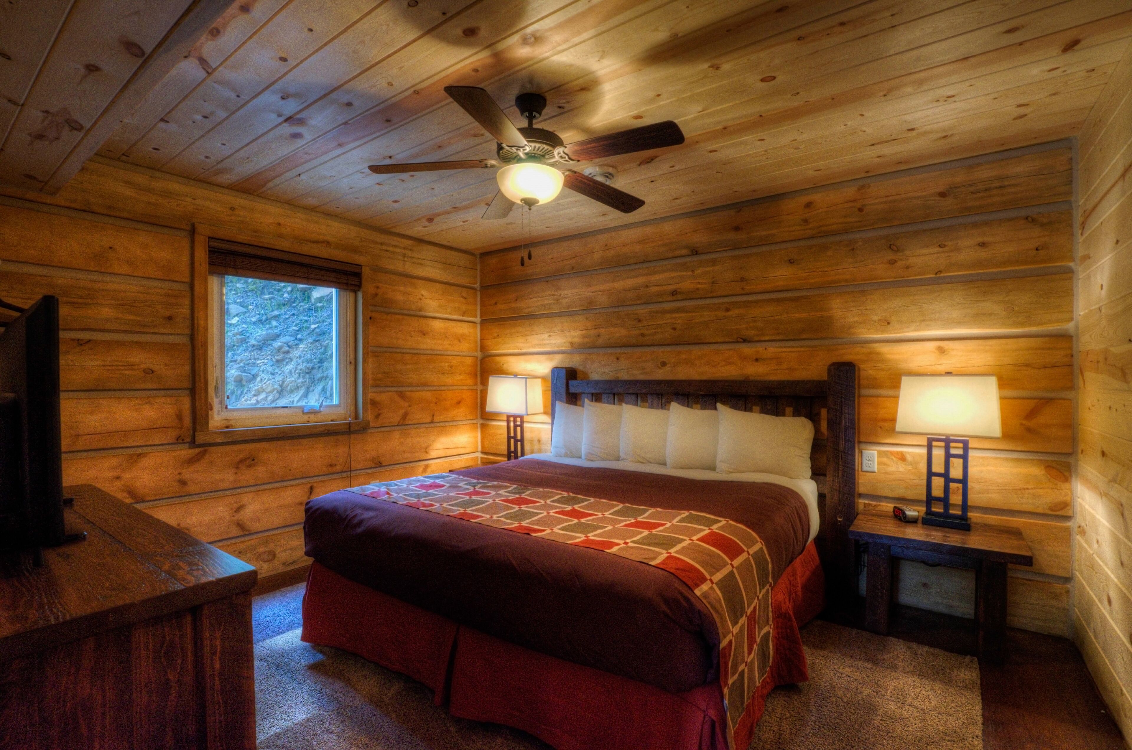 A wooden cabin room with a bed, two bedside tables with lamps, a ceiling fan, and a window.