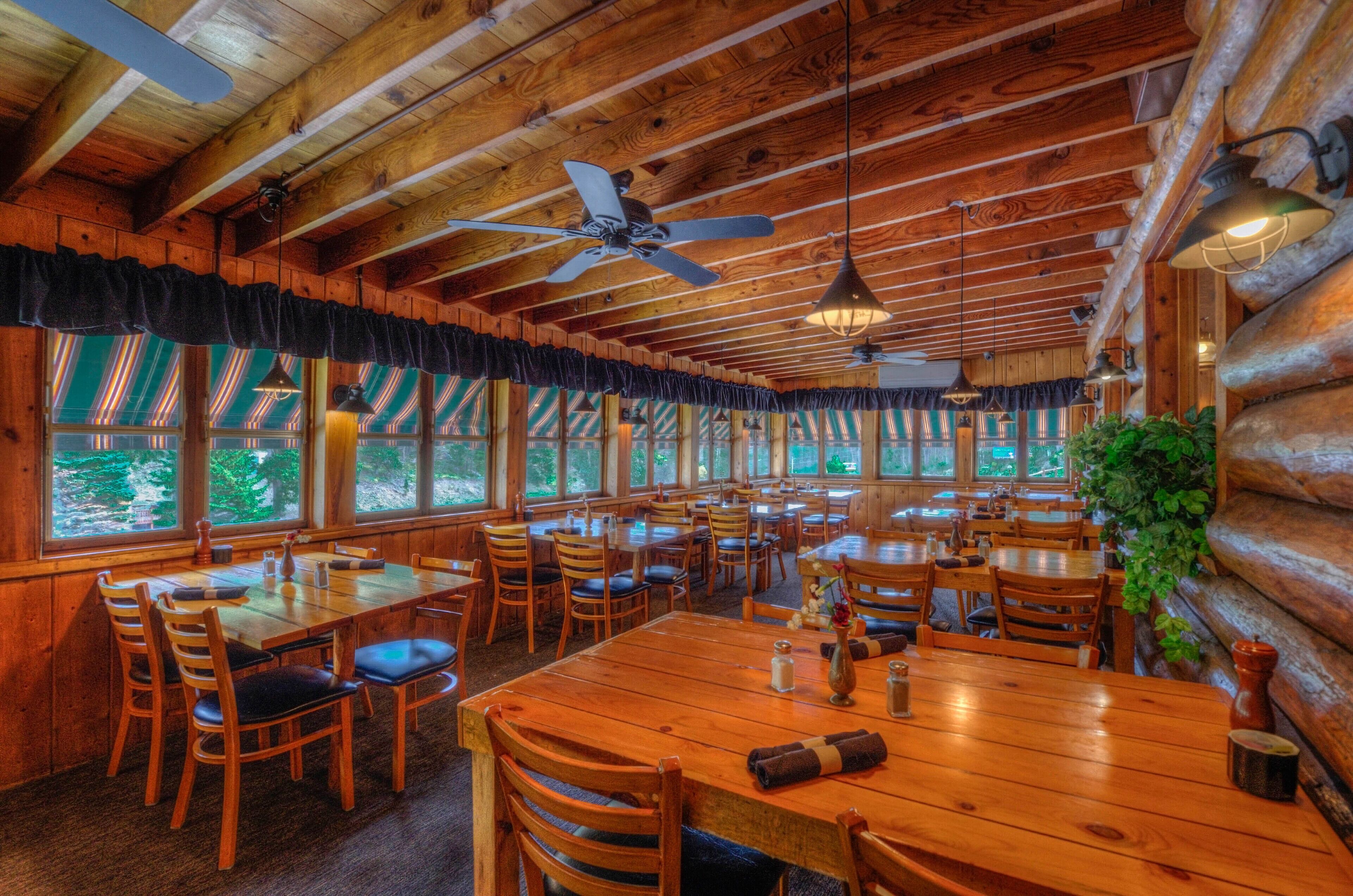 A log cabin-style restaurant with wooden tables and chairs, large windows, and a ceiling fan.