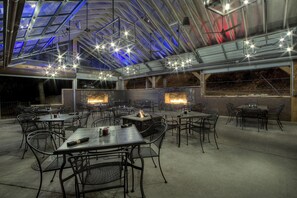 An outdoor dining area with tables and chairs, a fire feature, and a glass ceiling structure.