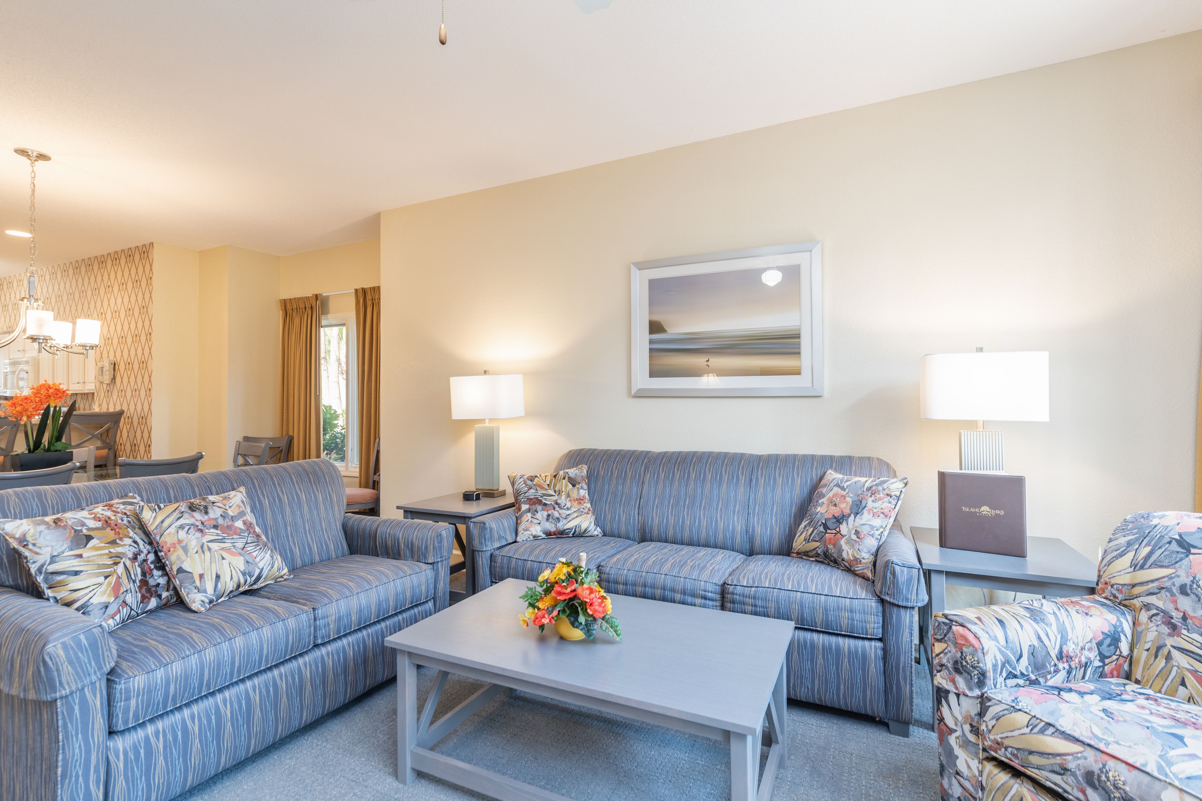 A living room with a sofa, armchair, coffee table, and lamps. There is a framed picture on the wall and a vase with flowers on the coffee table.