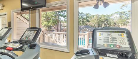A gym with treadmills and a heart rate monitor, large windows with a view of a pool and trees.