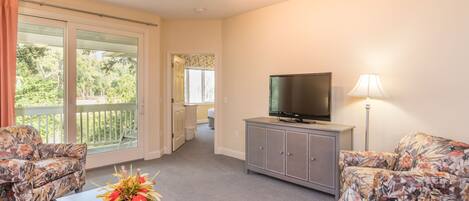 Een woonkamer met een flatscreen-televisie, sofa's met bloemenprint, een salontafel met een bloemstuk en een schuifpui die toegang geeft tot een balkon.