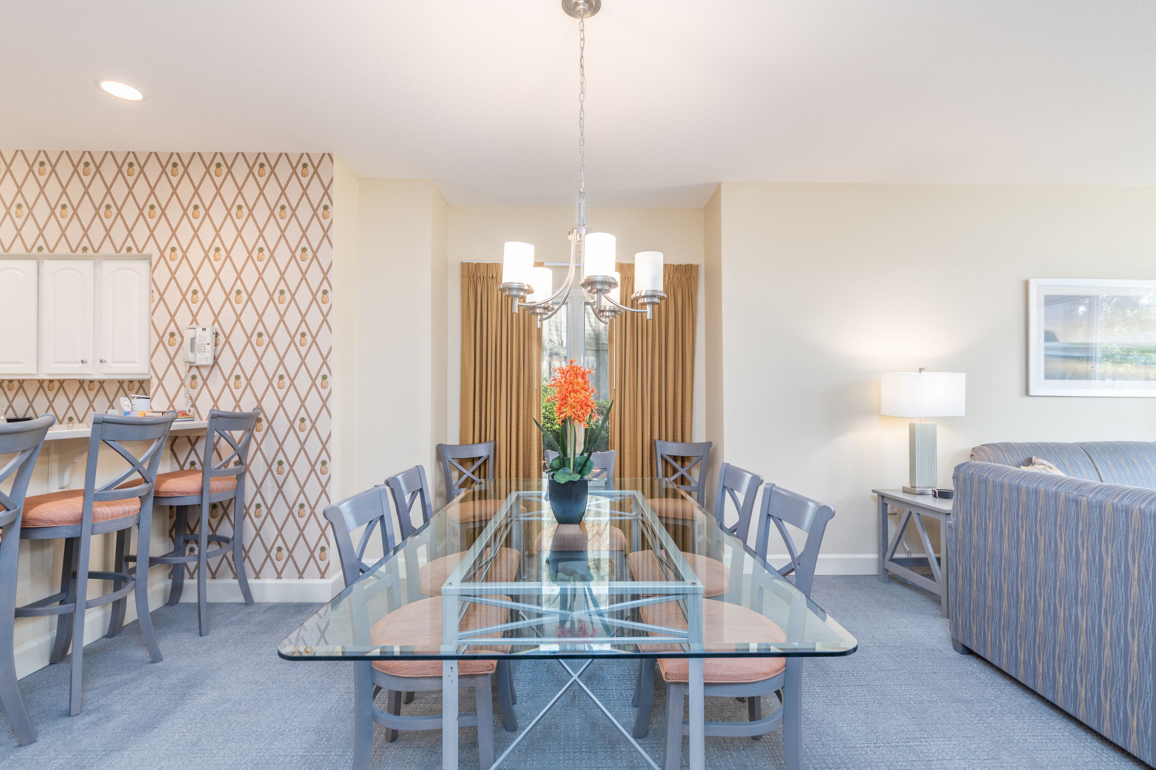 Une chambre d’hôtel avec une table à manger en verre, six chaises, un lustre, un canapé et une lampe.