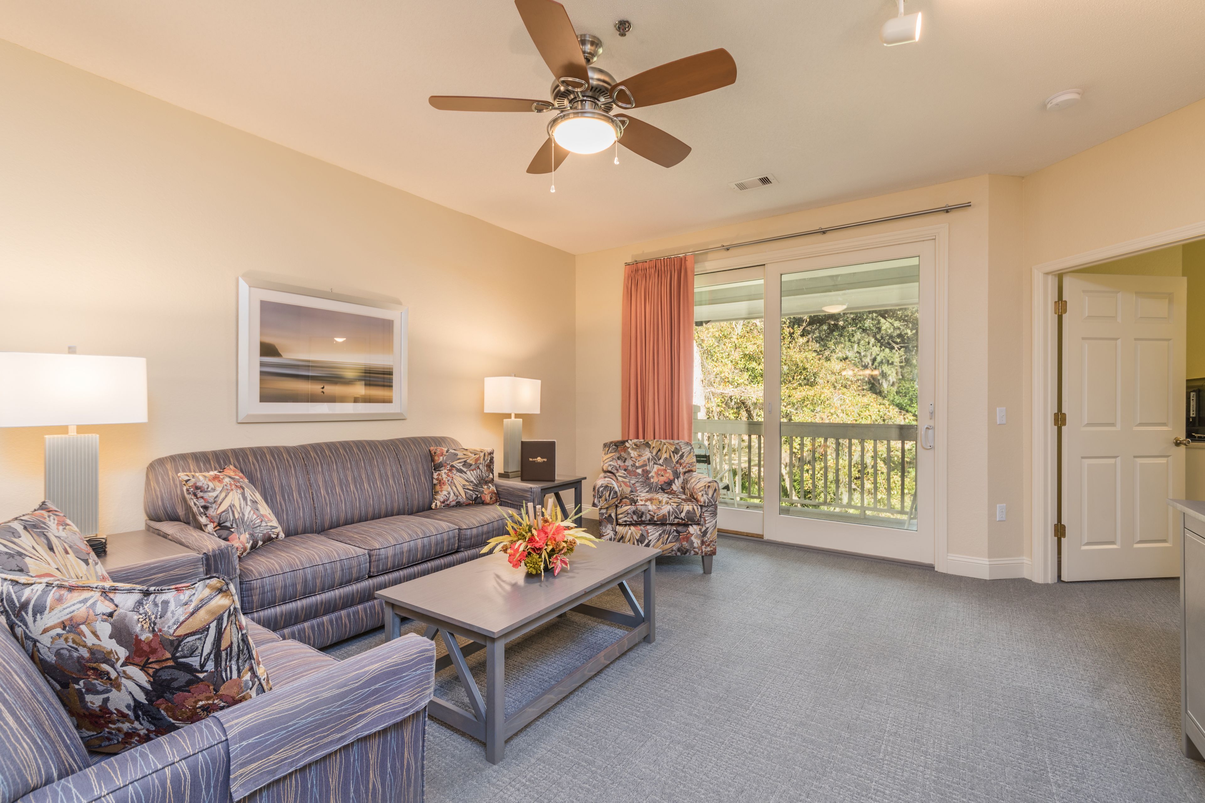 A living room with a sofa set, a coffee table, a ceiling fan, and a view of the outdoors through a sliding glass door.
