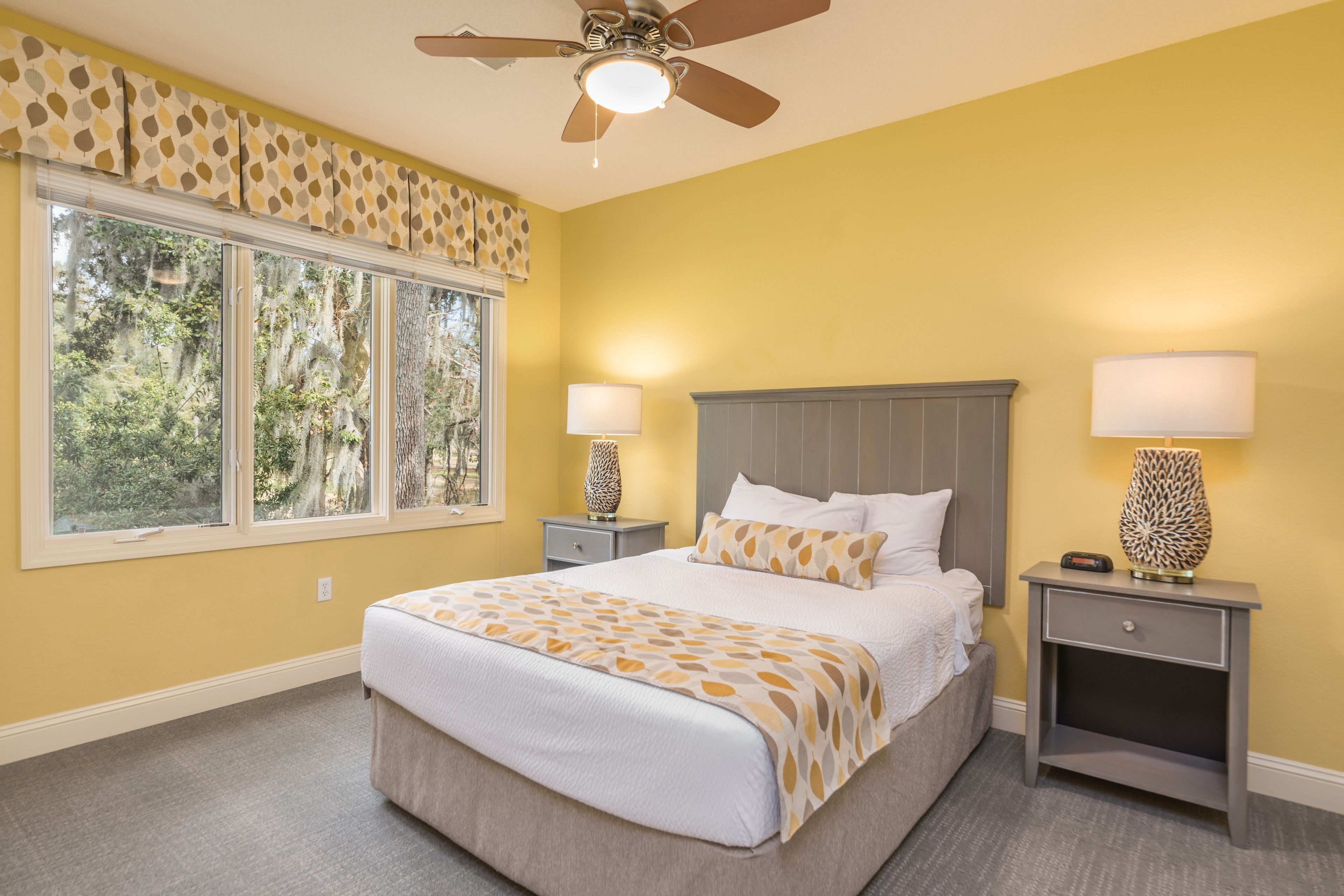 A bedroom with a large bed, two bedside tables with lamps, a ceiling fan, and a window with a view of trees.