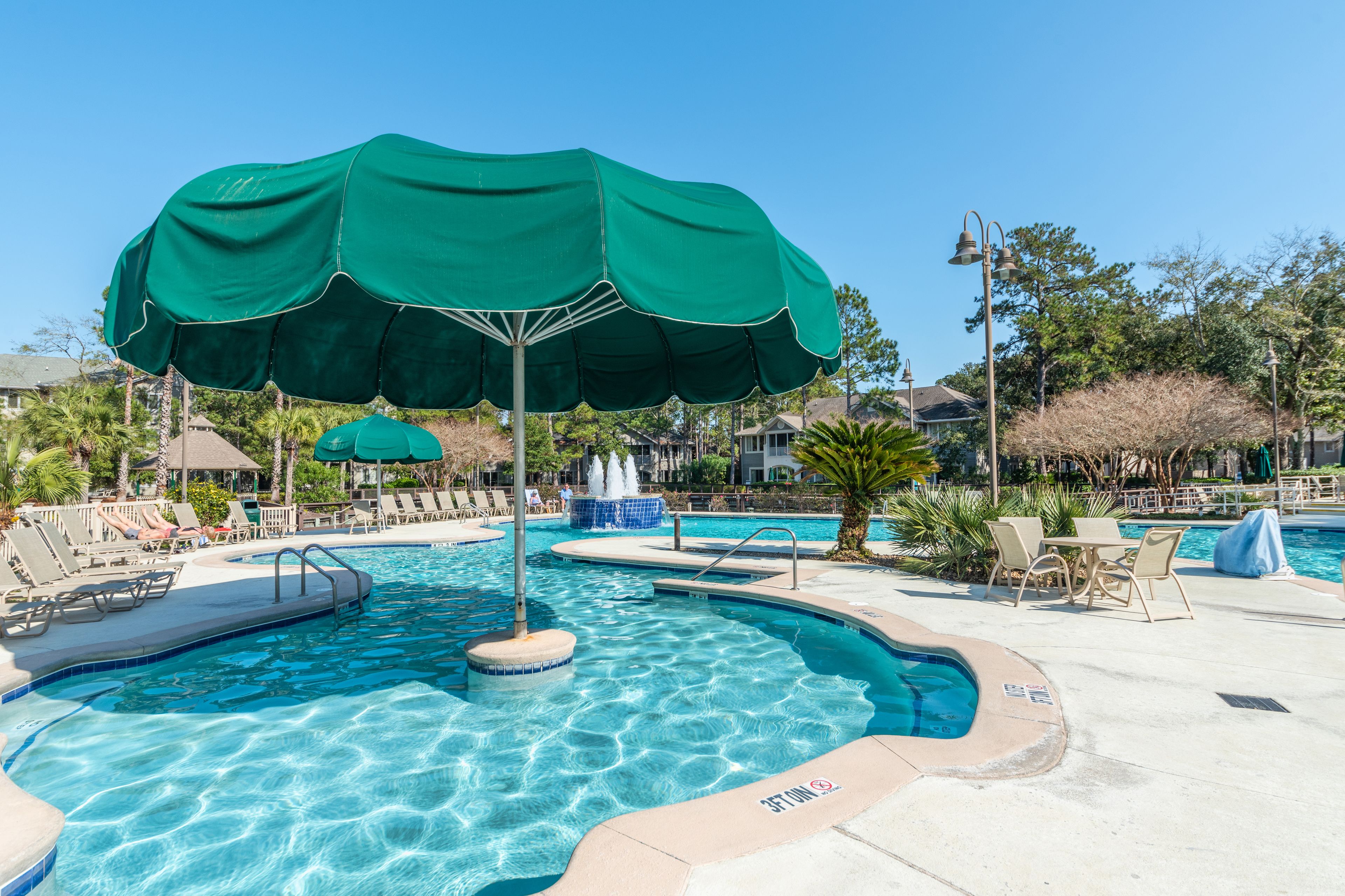 A swimming pool with lounge chairs and umbrellas, surrounded by palm trees and houses.