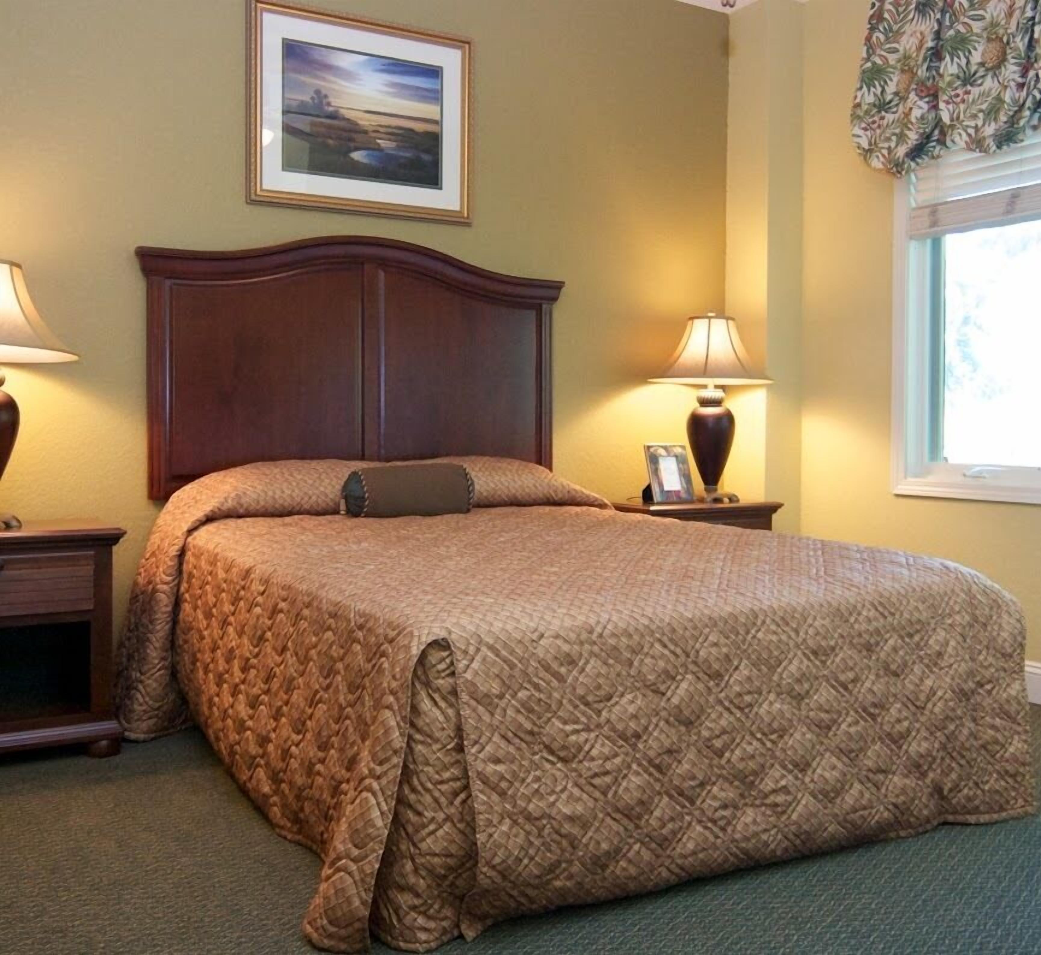 A neatly made bed with a quilted cover, two bedside tables with lamps, a framed picture on the wall, and a window with curtains.
