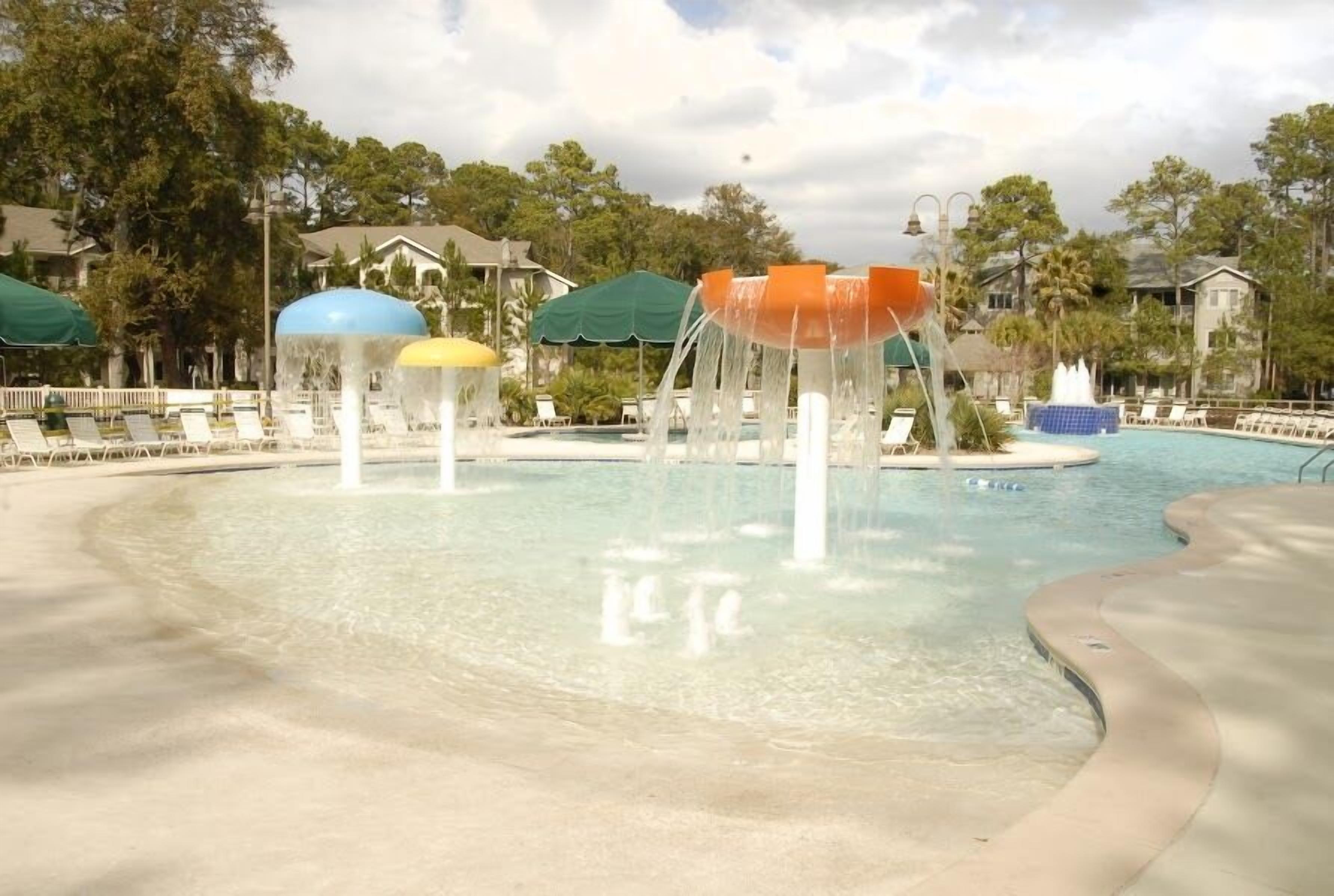 A pool area with multiple water features, including a central fountain and smaller water jets. There are lounge chairs and umbrellas around the pool, and a small island with a fountain.