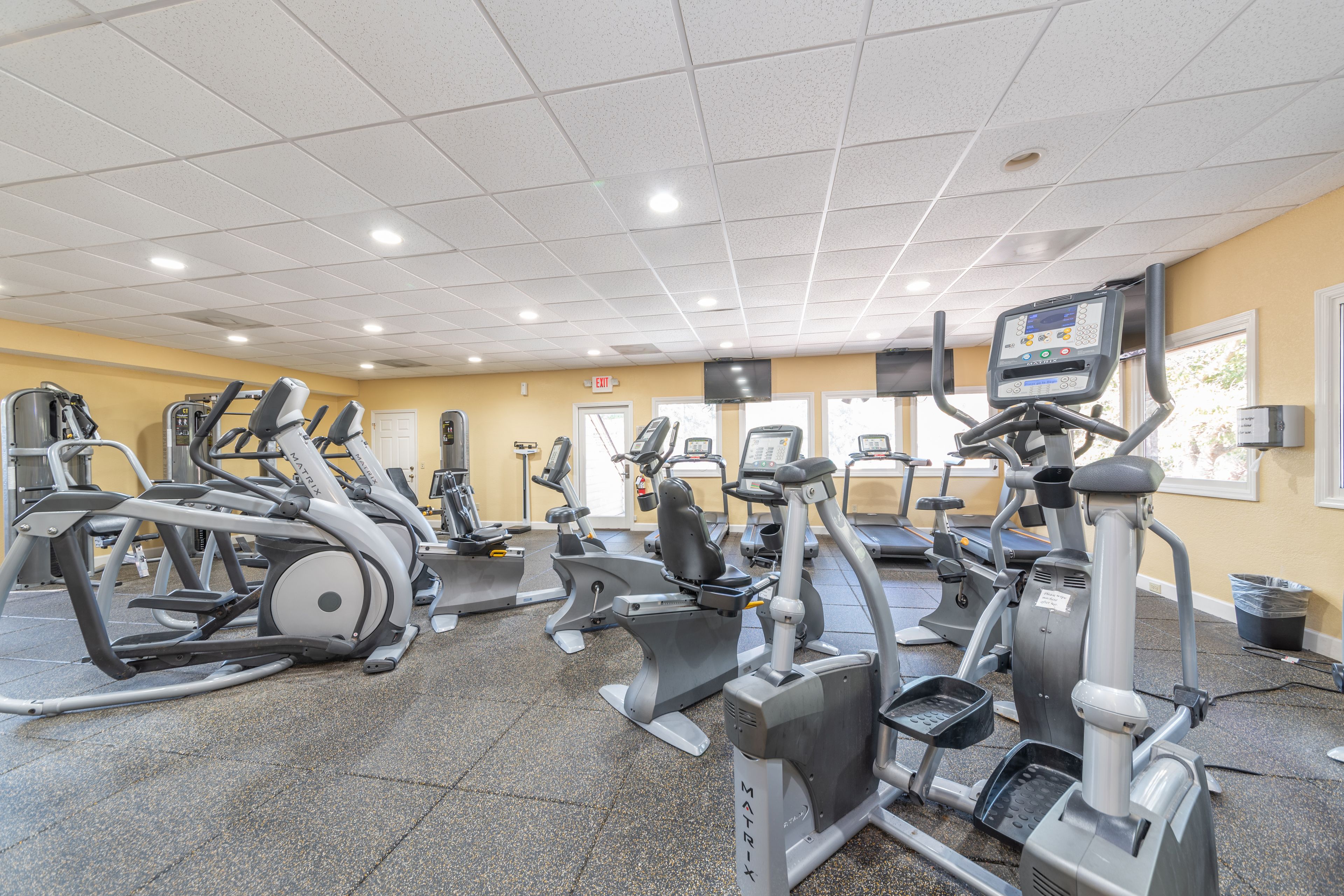 Un gimnasio con diversas máquinas de ejercicio, suelo de baldosas e iluminación empotrada en el techo.