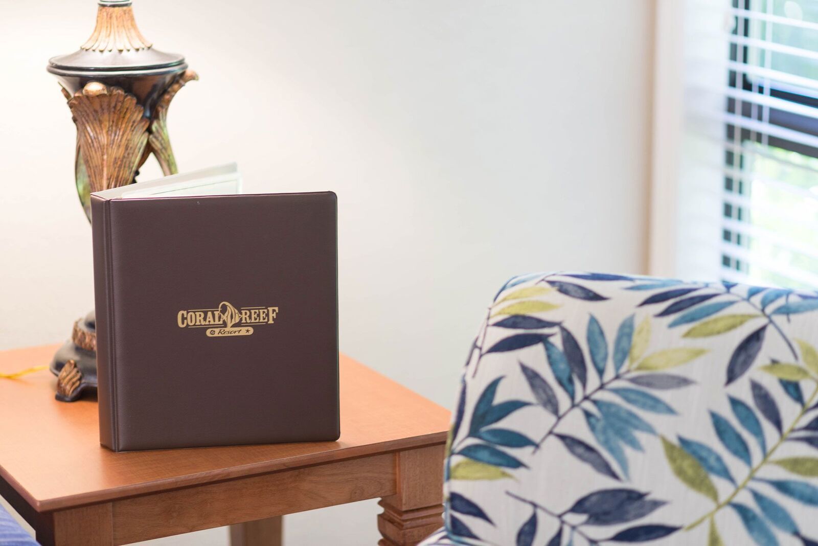 A dark-colored folder with "CORAL REEF Resort" branding on a wooden table next to a chair with a leaf-patterned cushion.