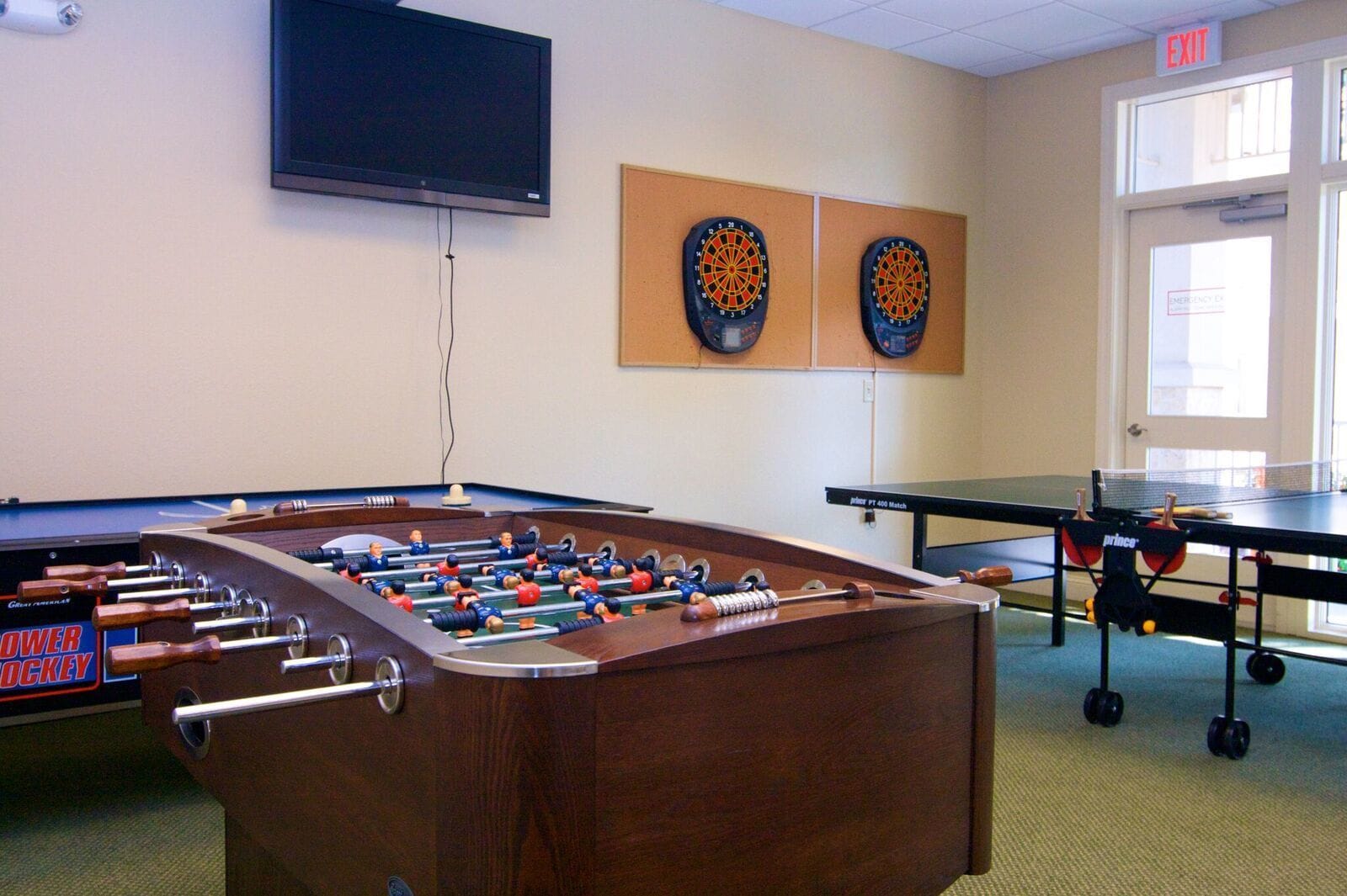A room with a foosball table, a ping pong table, and two dartboards on the wall.