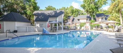 A swimming pool with lounge chairs and umbrellas, surrounded by houses with a covered patio area.