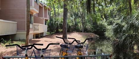 A row of bicycles with baskets, a building with a balcony, and a pond surrounded by trees.