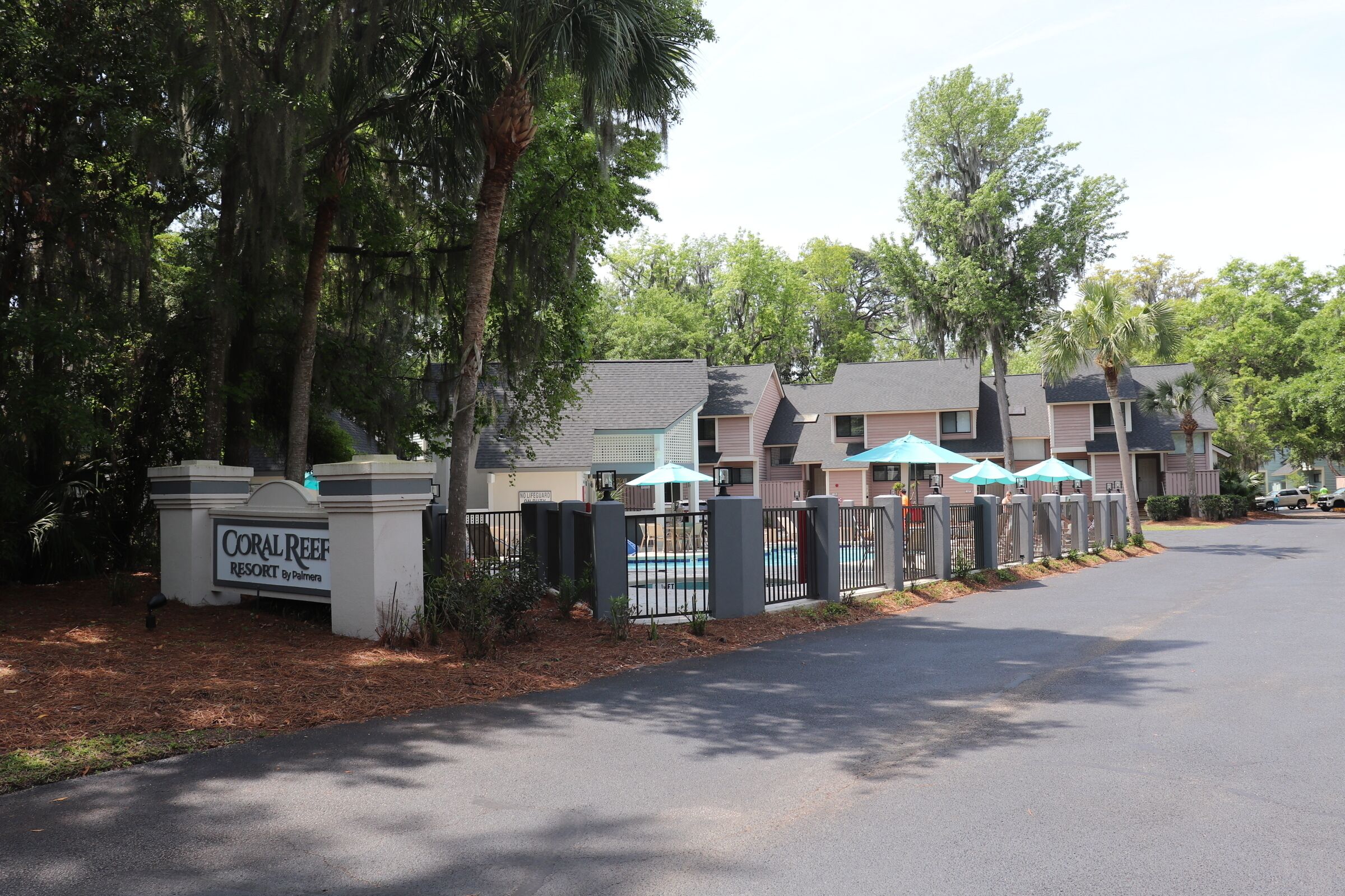 A resort entrance with a gated driveway, palm trees, and a sign for Coral Reef Resort by Palmira.