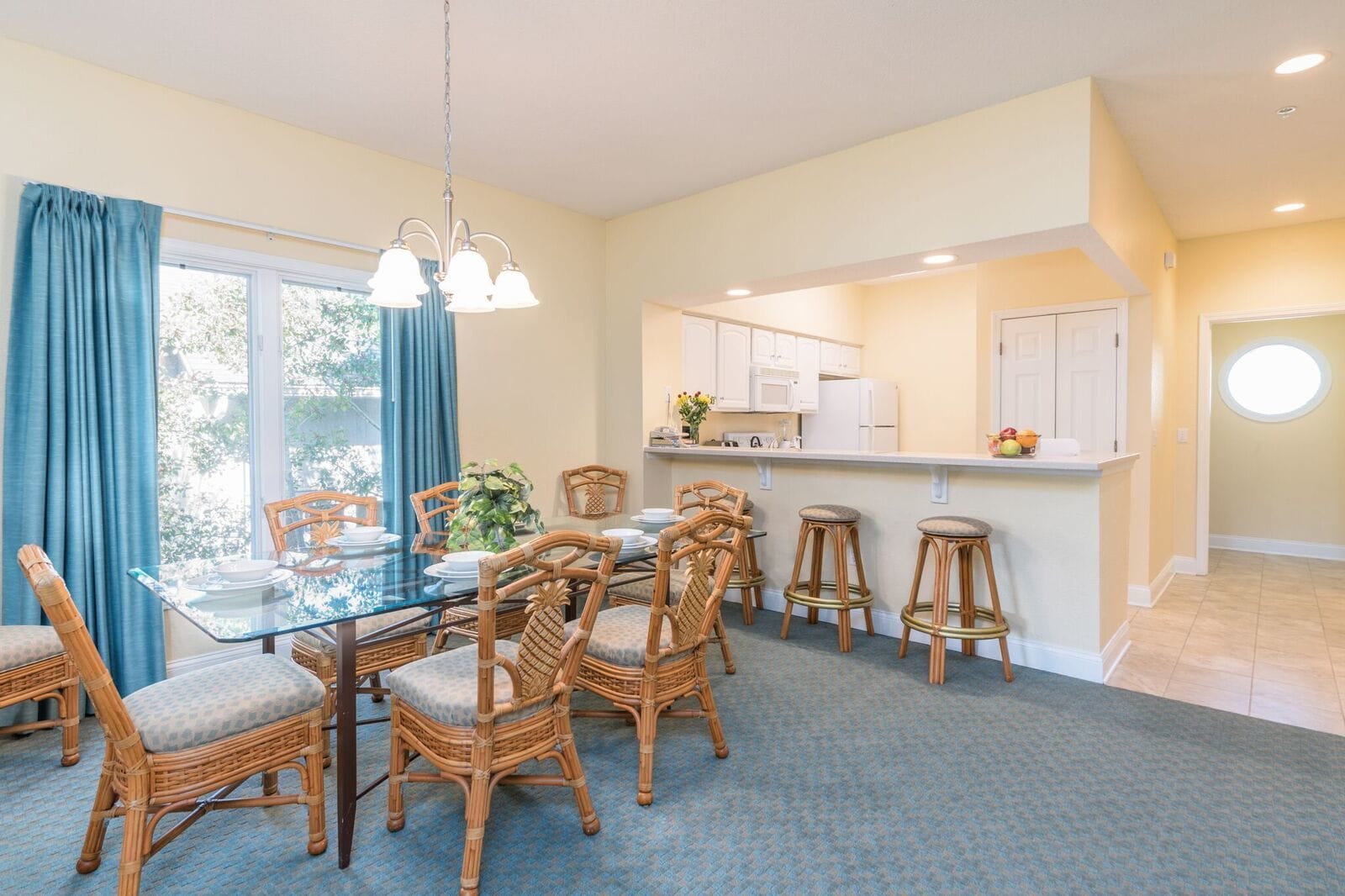 A dining area with a glass table, wicker chairs, and a kitchen in the background.