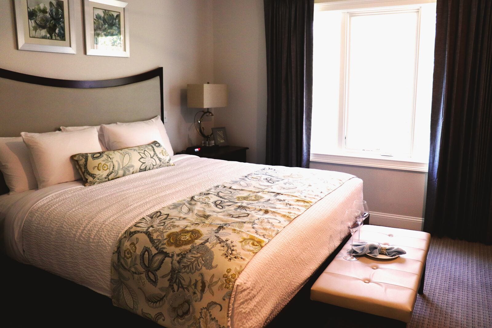 Un lit bien fait, recouvert d’une couvre-lit à motifs floraux, une table de chevet avec une lampe et une fenêtre avec des rideaux.