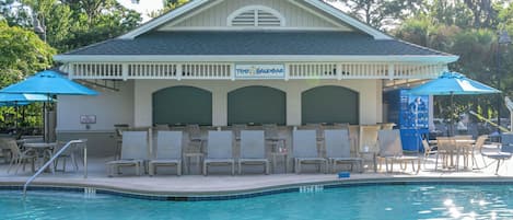 A pool area with lounge chairs, umbrellas, and a building with a sign reading 'The Sandbar.'