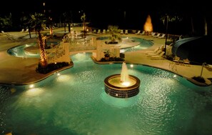 A nighttime view of a resort pool area with multiple pools, a central fountain, and surrounding lounge chairs.