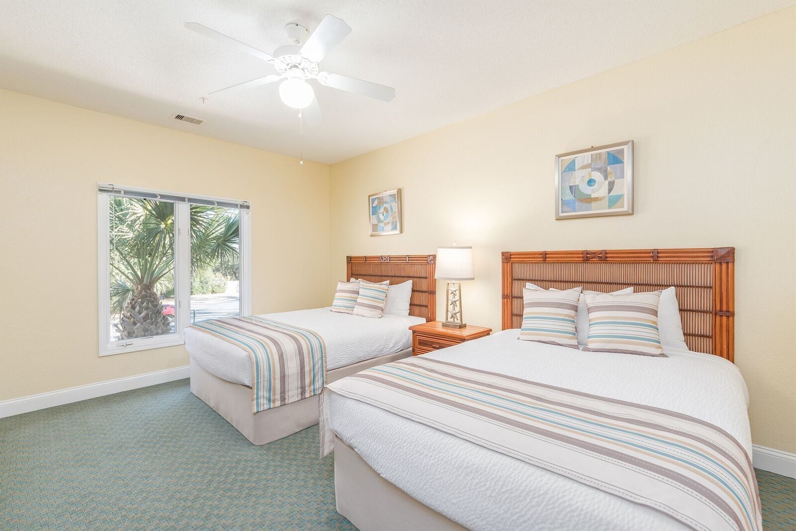 A hotel room with two beds, a ceiling fan, a window with a view of palm trees, and artwork on the walls.