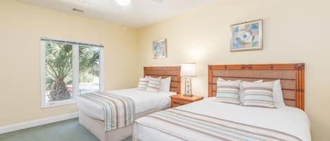 A hotel room with two beds, a ceiling fan, a window with a view of palm trees, and artwork on the walls.