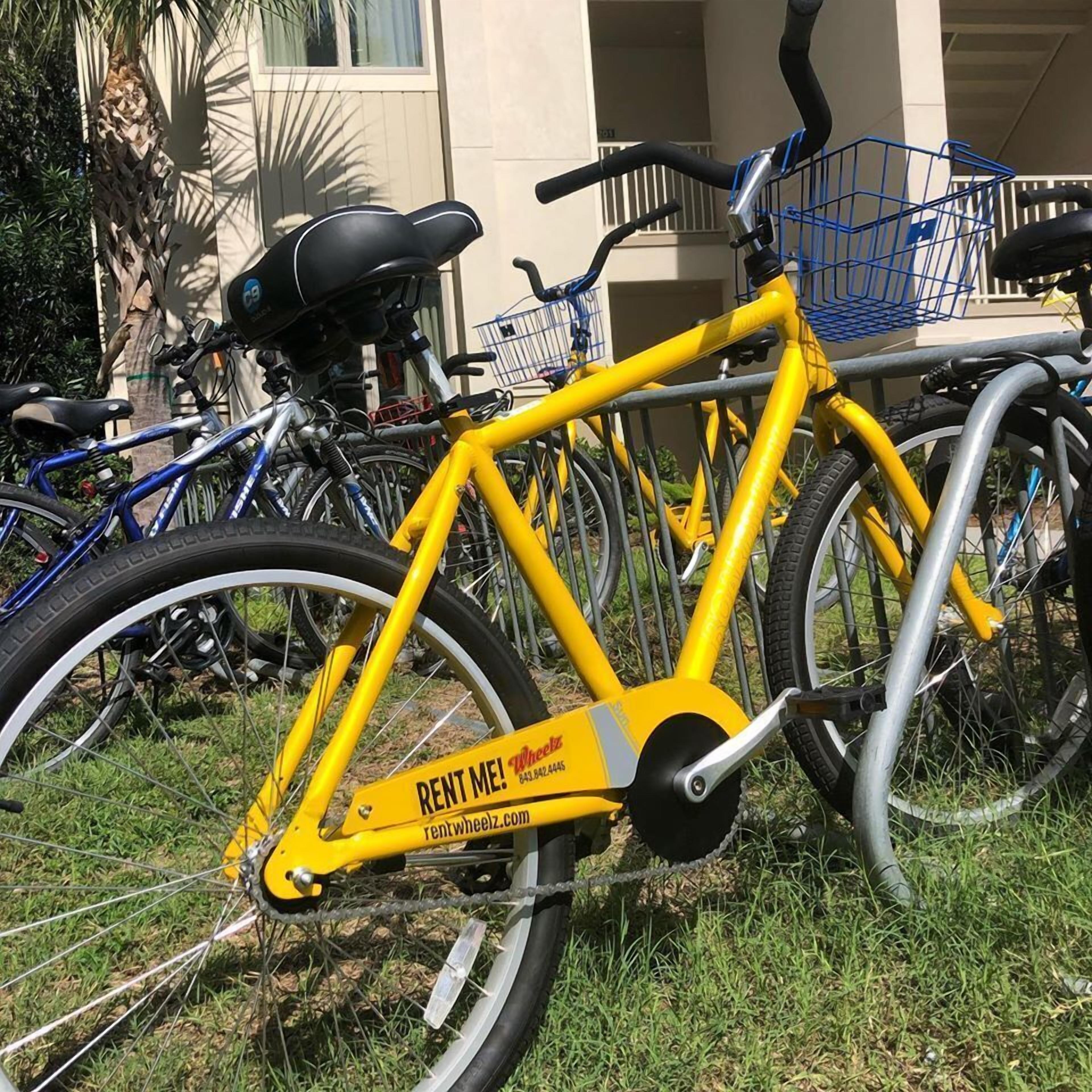 A yellow bicycle with a 'RENT ME!' sign, parked on grass with other bicycles in the background.