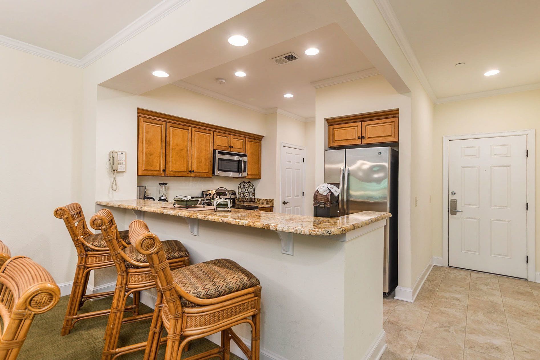 A kitchen with wooden cabinets, a granite countertop, a refrigerator, and wicker chairs.