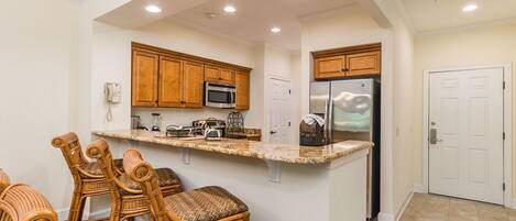 A kitchen with wooden cabinets, a granite countertop, a refrigerator, and wicker chairs.