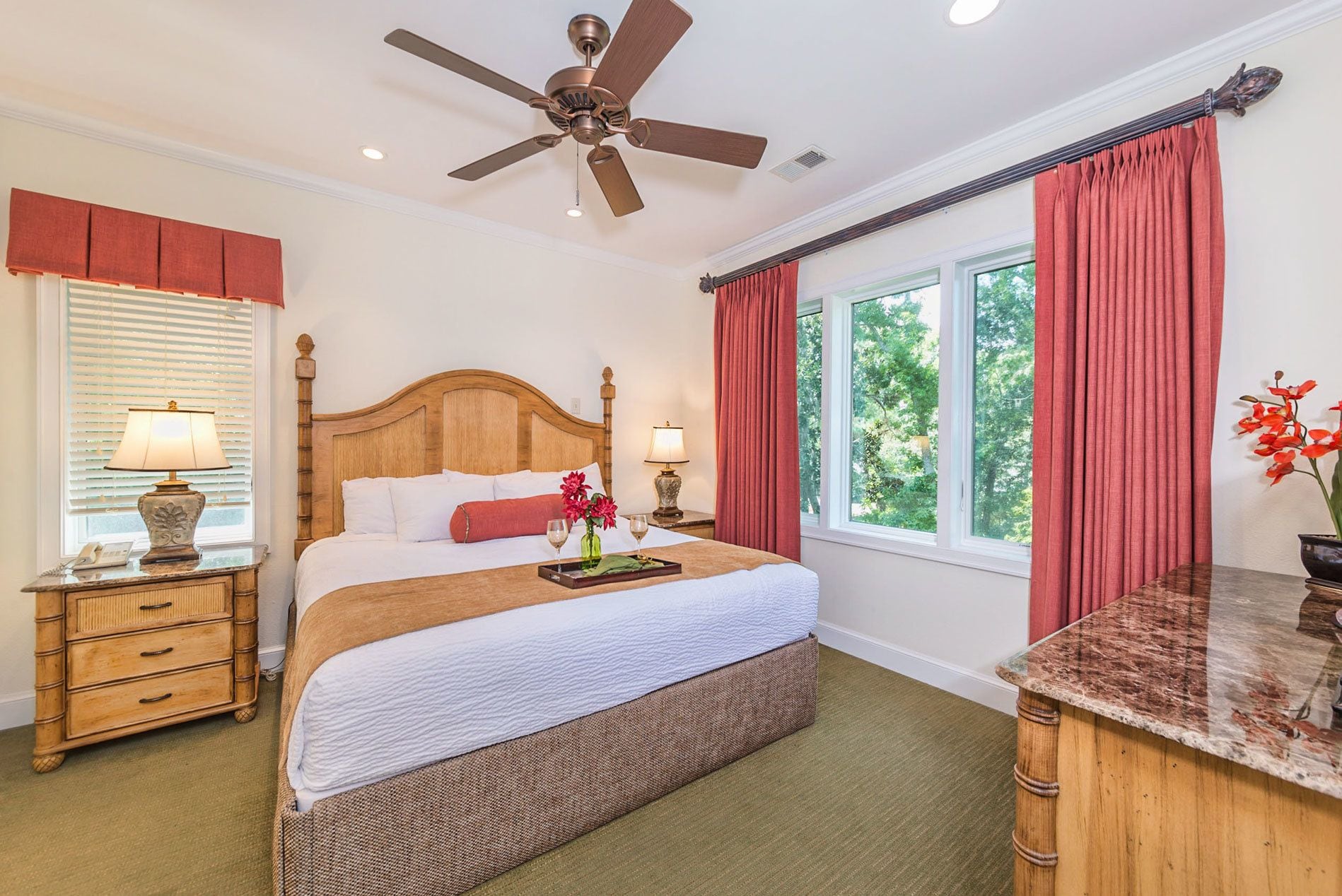 A bedroom with a wooden bed, two bedside tables with lamps, a ceiling fan, and a window with red curtains.