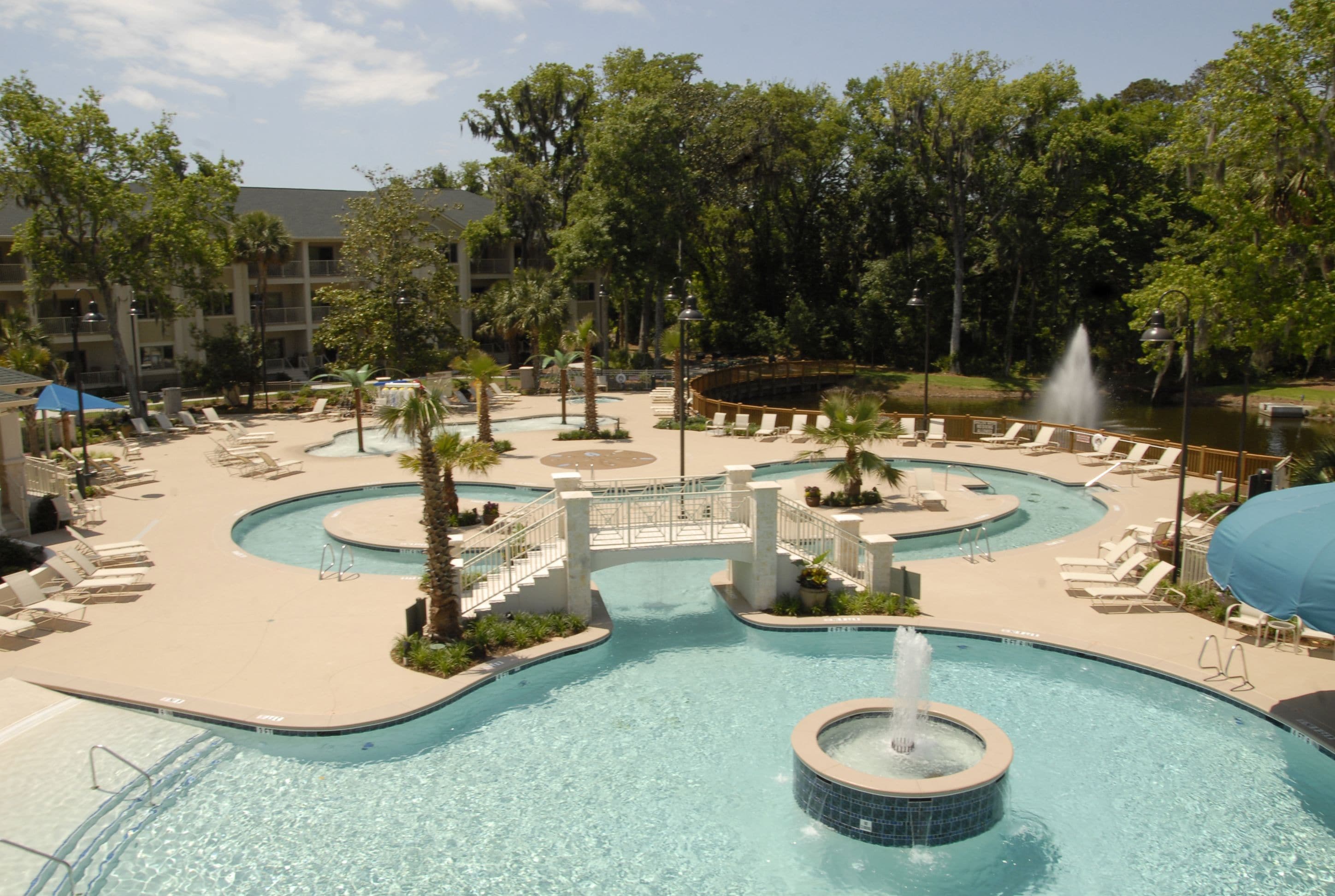 A pool area with multiple water features, including a central fountain, surrounded by lounge chairs and palm trees.