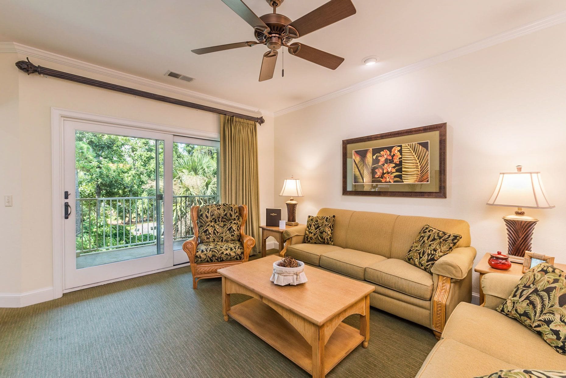A living room with a sofa, armchairs, a coffee table, a ceiling fan, and a large window with a view of greenery.