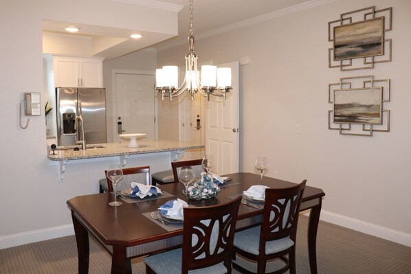 A dining area with a wooden table, chairs, a chandelier, and a kitchen in the background.