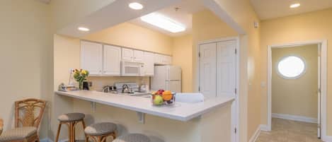 A kitchen with white cabinets, a central island with stools, a microwave, and a fruit bowl.