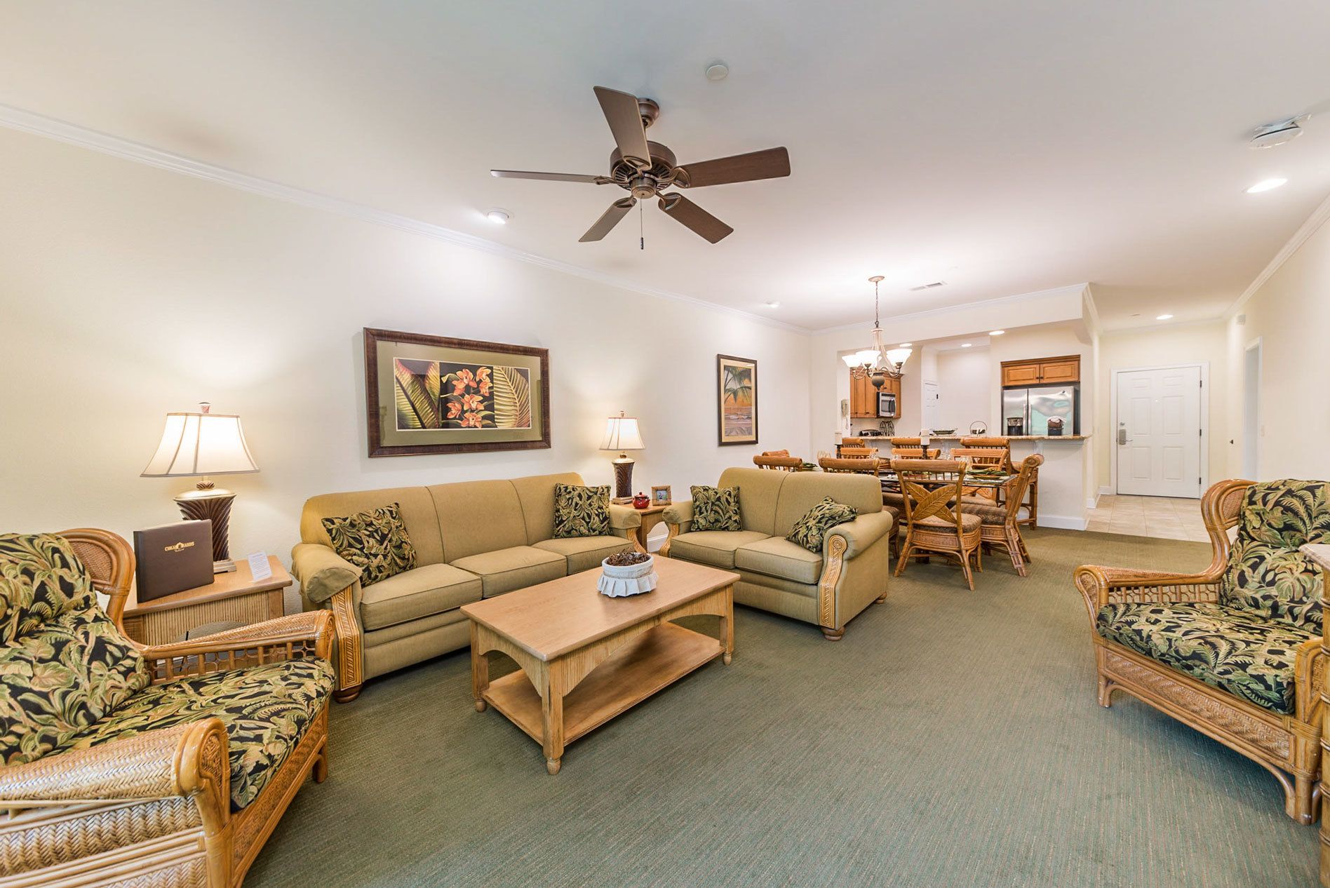 A spacious living room with a sofa, chairs, a coffee table, and a dining area with a table and chairs. There is a ceiling fan, a painting on the wall, and a kitchen area in the background.