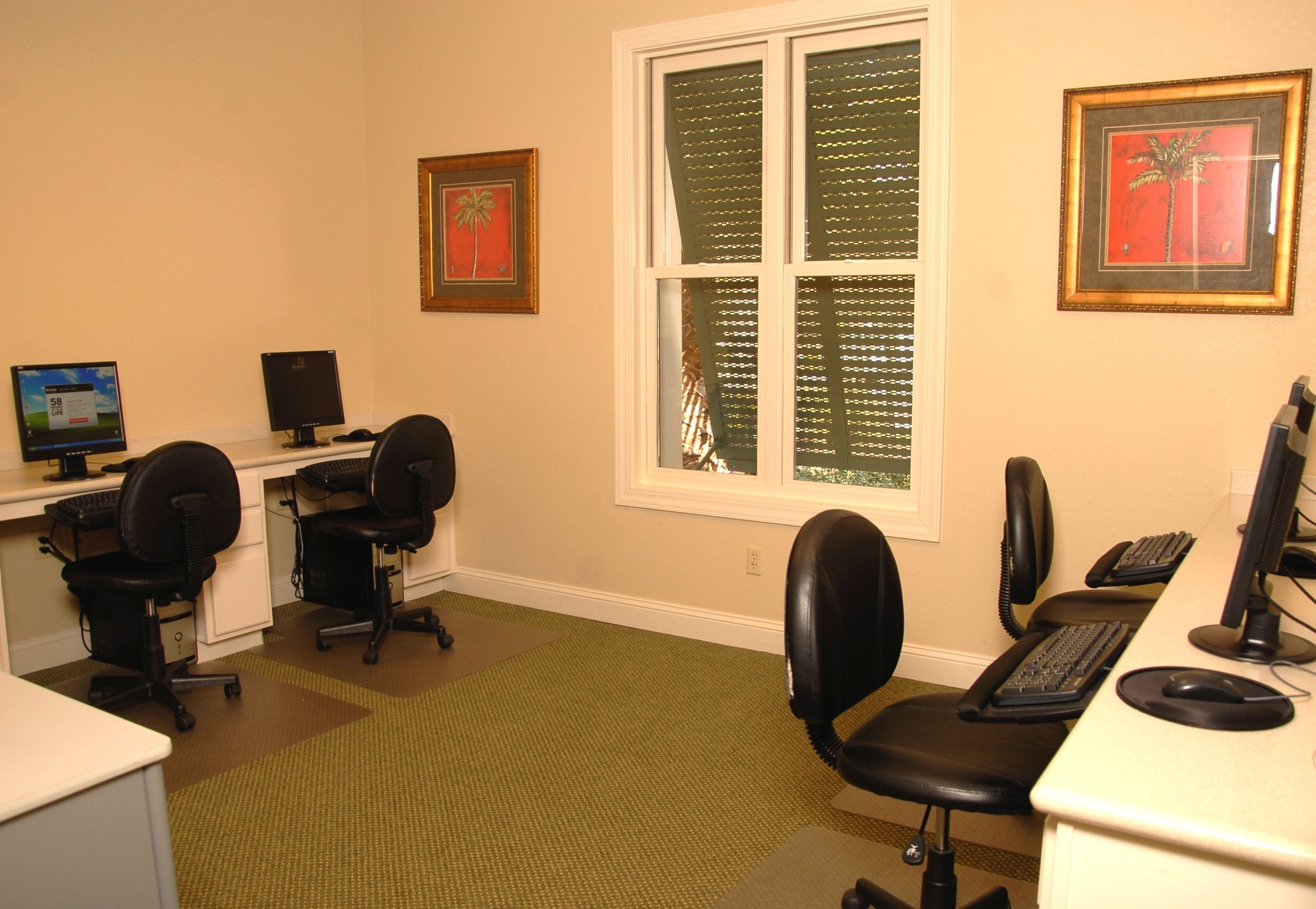 A room with two desks, two chairs, a window with blinds, and framed pictures on the wall.