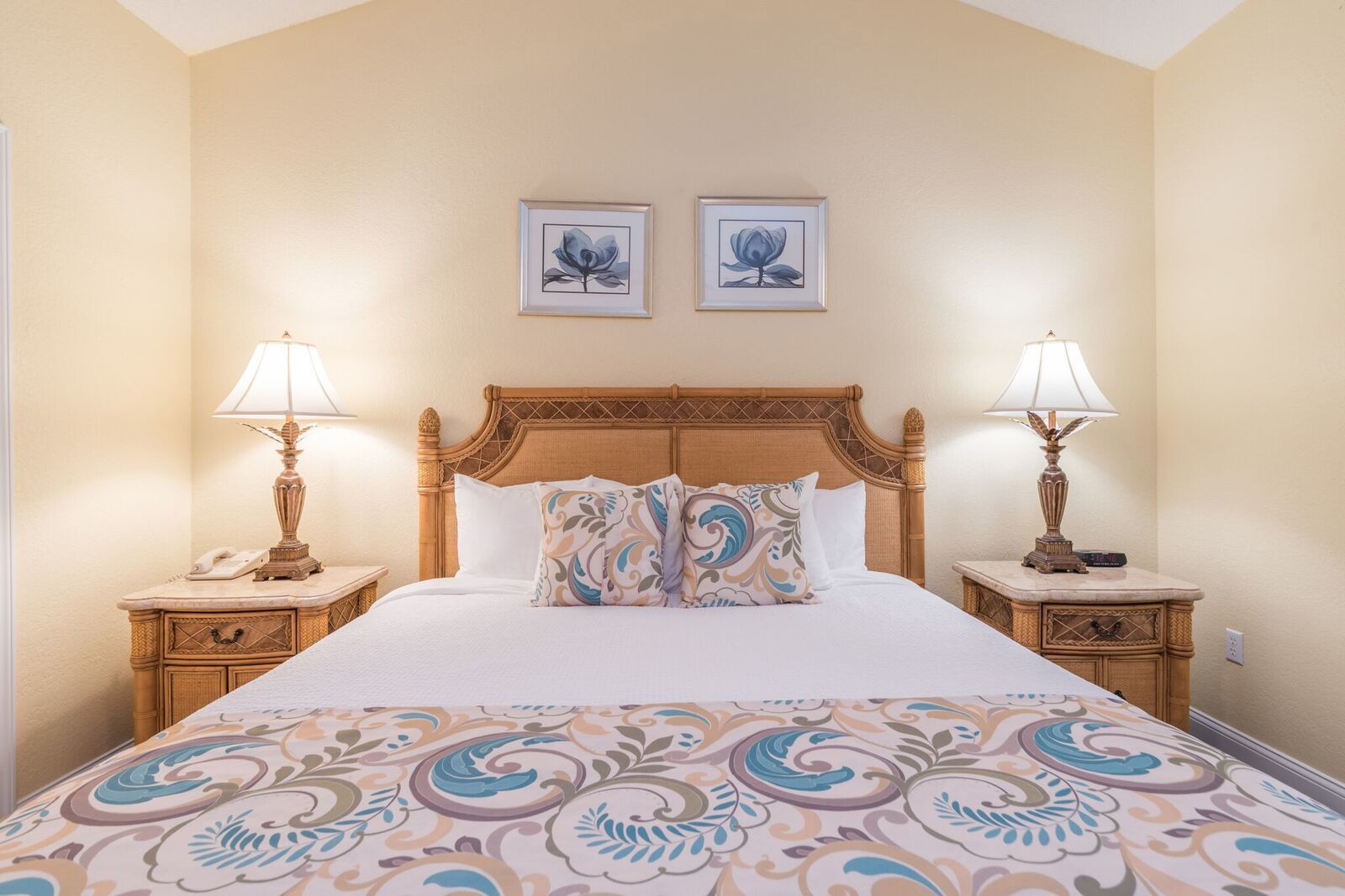 Une chambre à coucher avec un grand lit, deux tables de chevet avec des lampes, une tête de lit et deux tableaux floraux encadrés accrochés au mur.