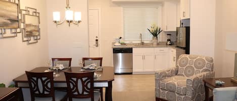 Un comedor con una mesa y sillas de madera, una cocina con armarios blancos, un sillón estampado y una mesita auxiliar con un florero.