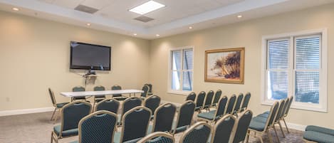 A conference room with a large table, rows of chairs, a flat-screen TV, windows with blinds, and a framed artwork on the wall.